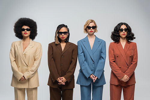 fashionable multiracial female friends in dark sunglasses and multicolored suits on grey backdrop