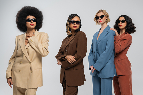 group of trendy multiracial girlfriends in dark sunglasses and multicolored suits on grey, fashion