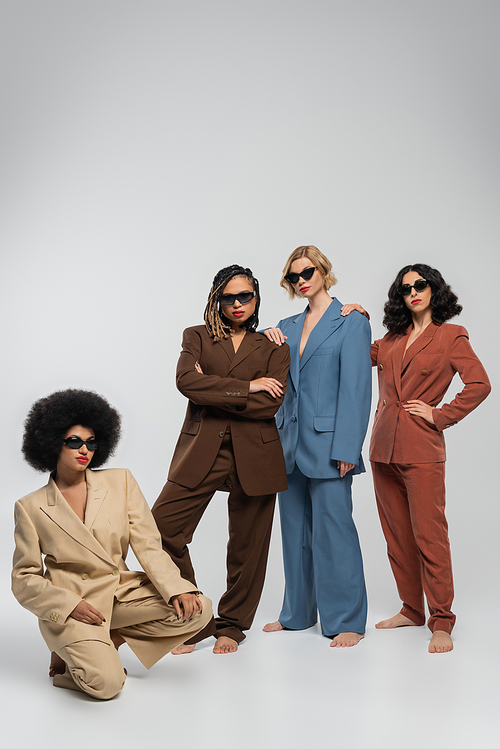 barefoot multiethnic fashion models in dark sunglasses and colorful suits on grey, full length