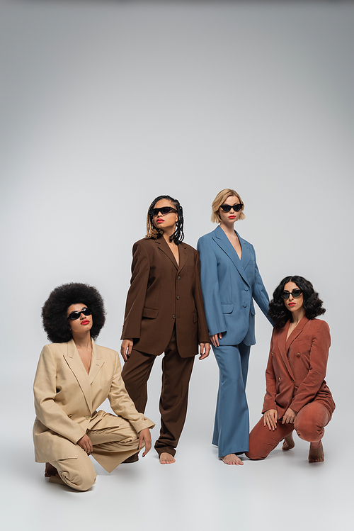 full length of fashionable multiracial women in colorful suits and dark sunglasses on grey backdrop