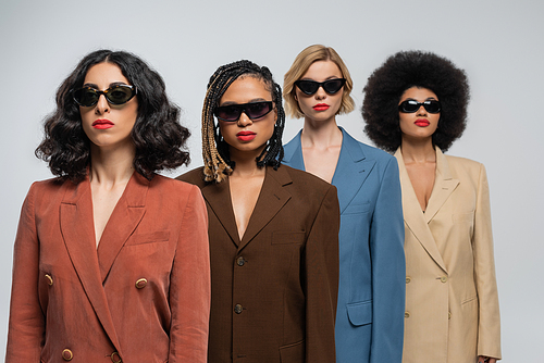 diverse group of fashionable multiracial girlfriends in dark sunglasses and colorful suits on grey