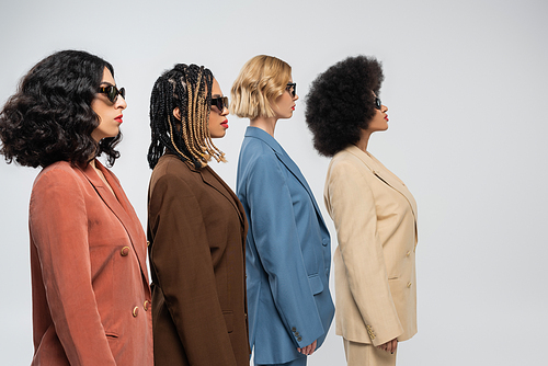 side view of stylish multiracial girlfriends in dark sunglasses and multicolored suits on grey