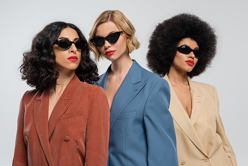 three fashionable multiracial girlfriends in multicolored suits and dark sunglasses on grey