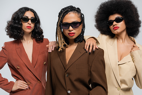 multiracial women in sunglasses and suits touching shoulders of african american girlfriend on grey