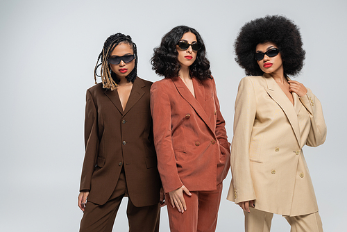 glamorous multiethnic girlfriends in colorful suits and dark sunglasses posing on grey, diversity