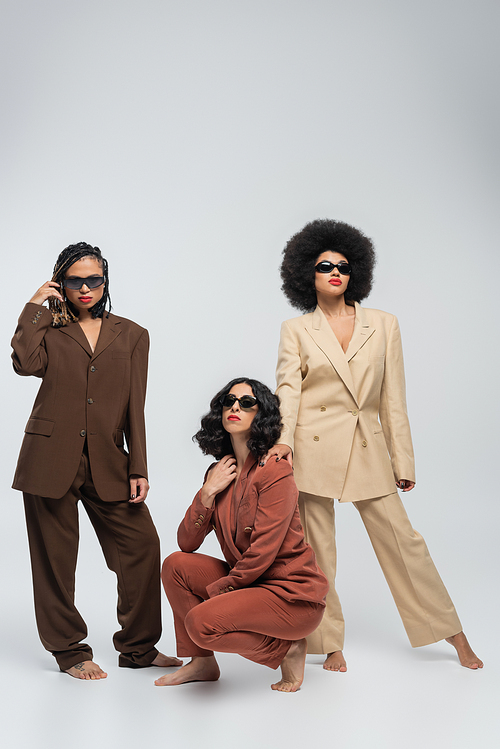 barefoot multiracial female model in trendy colorful suits and dark sunglasses on grey, full length