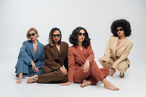 diverse group of stylish multiracial women in dark sunglasses and colorful suits sitting on grey