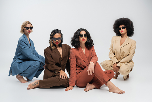 diverse fashion, multiracial girlfriends in dark sunglasses and multicolored suits sitting on grey