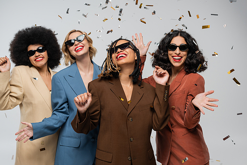 overjoyed multiracial girlfriends in sunglasses and trendy suits under colorful confetti on grey
