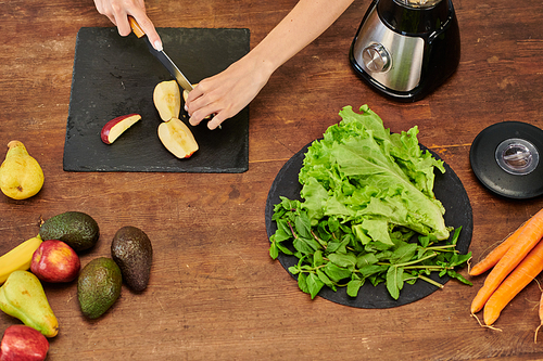 cropped view of woman cutting apple near electric blender and lettuce with mint, plant-based diet