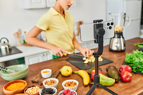 selective focus of smartphone on tripod near vegetarian video blogger cutting fruits in kitchen