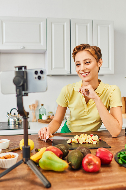 happy vegetarian woman looking at smartphone on tripod near fresh fruits in kitchen, culinary vlog