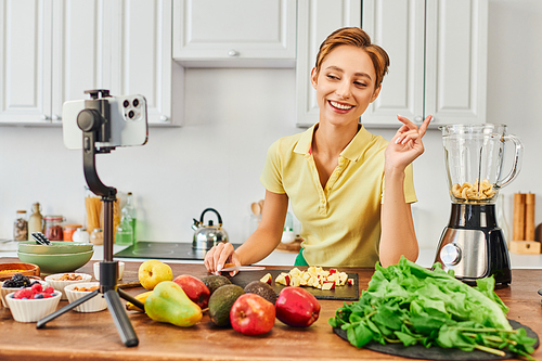 cheerful vegetarian woman looking at smartphone on tripod near fruits and electric blender, vlog