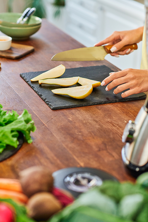 partial view of vegetarian woman with knife near ripe sliced pear on chopping board in kitchen