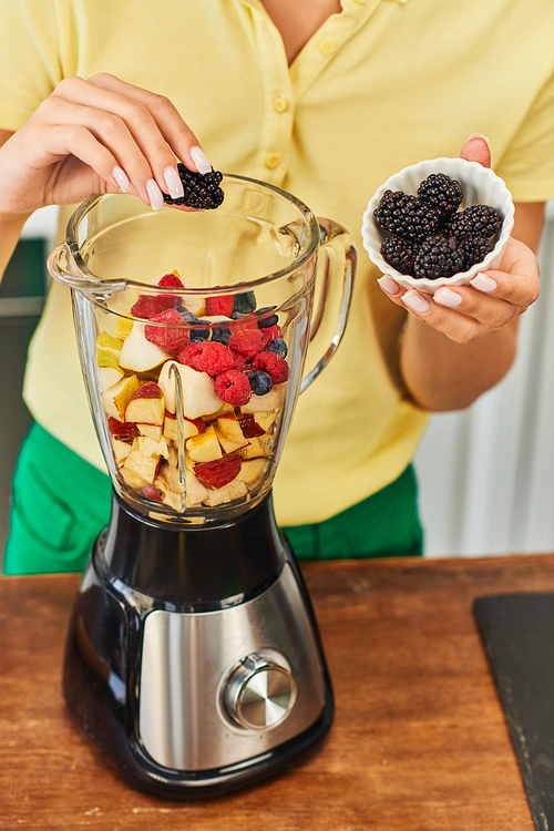 cropped view of vegetarian woman adding blackberries into electric blender with delicious fruits