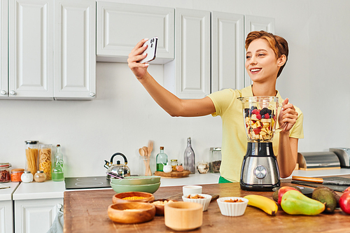 pleased vegetarian woman taking selfie on smartphone near electric blender with fresh chopped fruits