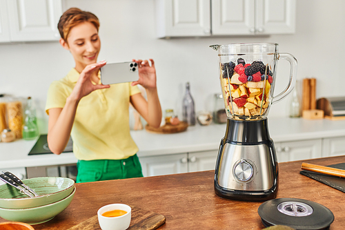 happy woman with smartphone taking photo of electric blender with chopped fruits, vegetarian recipe