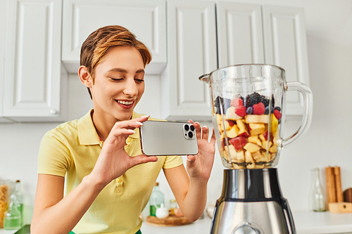 positive woman taking photo of electric blender with chopped fruits, delicious plant-based diet