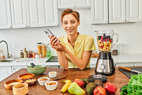 vegetarian woman with smartphone smiling at camera near plant origin food and blender with fruits