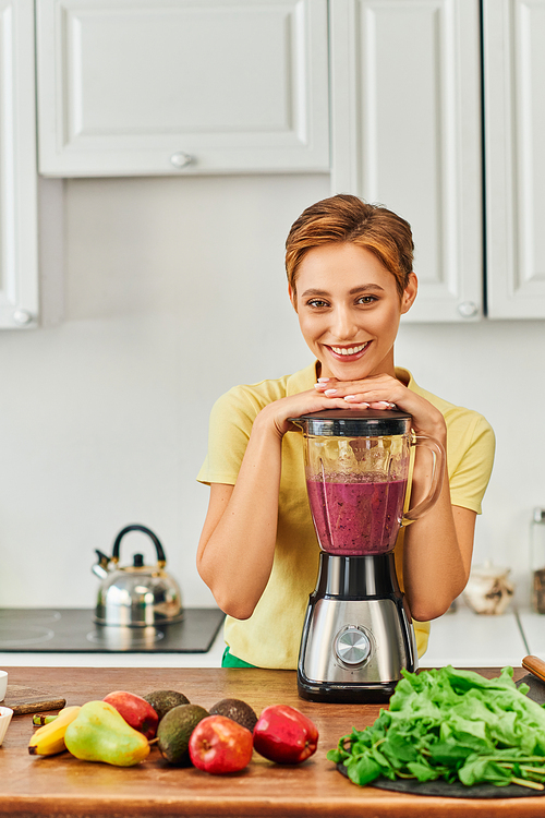 vegetarian woman smiling at camera near blender with wholesome smoothie in kitchen, plant-based diet