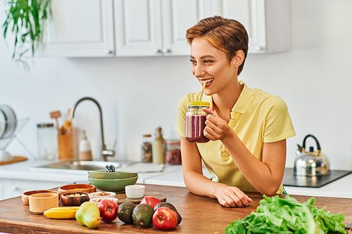 smiling vegetarian woman drinking delicious smoothie from mason jar with straw in kitchen
