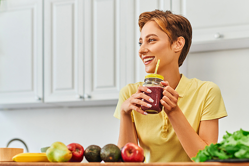 joyful vegetarian woman holding fresh smoothie in mason jar with straw and looking away in kitchen