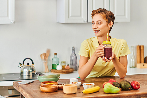 smiling woman holding mason jar with plant-based smoothie near fresh fruits on table in kitchen