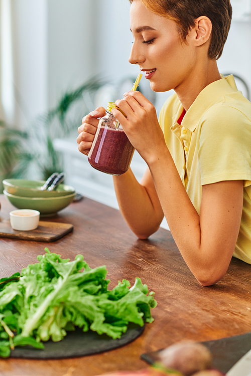 young vegetarian woman holding mason jar with delicious smoothie near fresh lettuce on kitchen table