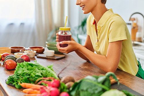 cropped woman with vegetarian smoothie in mason jar near plant-based ingredients on kitchen table