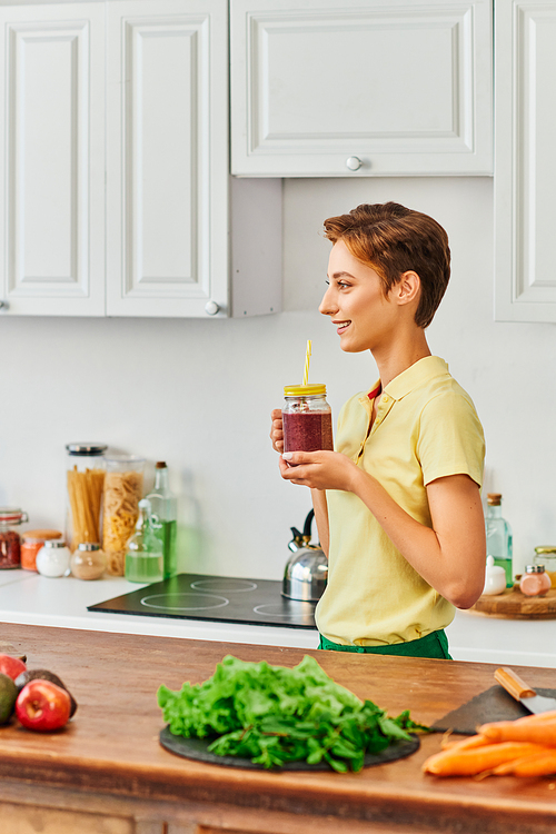 side view of smiley vegetarian woman holding mason jar with fresh smoothie in modern kitchen