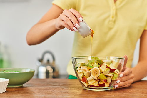 cropped view of woman pouring honey into bowl with chopped fruits, wholesome vegetarian salad