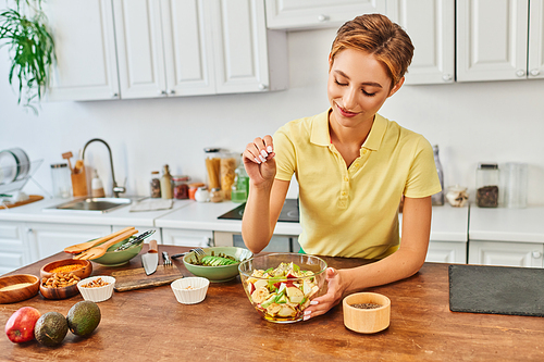 smiling woman seasoning fresh fruit salad with sesame seeds in kitchen, delicious vegetarian diet