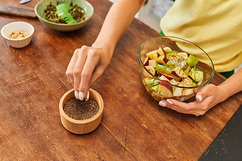 cropped view of woman taking sesame seeds from wooden bowl near fresh fruit salad, vegetarian diet