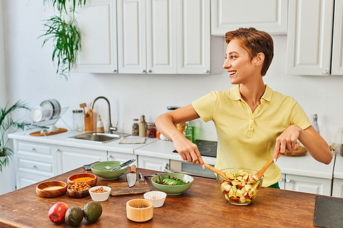 cheerful woman mixing vegetarian salad bear fresh fruits and looking away in modern kitchen