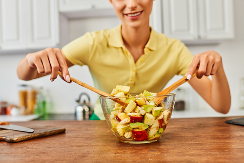 partial view of woman mixing fresh fruit salad with wooden spatulas in kitchen, vegetarian diet