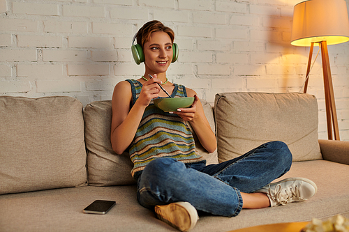 smiling woman in headphones with salad bowl on couch in living room, vegetarian evening snack