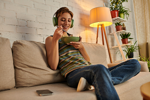cheerful woman in headphones having vegetarian evening snack on couch in cozy living room