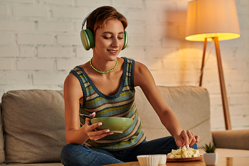smiley woman in headphones holding salad bowl and taking tofu cheese, vegetarian dinner on couch