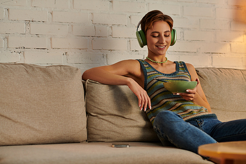 pleased veggie woman in headphones sitting with salad bowl on couch in cozy evening living room