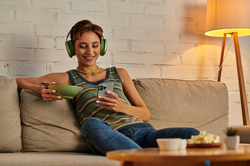 happy veggie woman in headphones networking on smartphone while sitting with salad bowl on couch