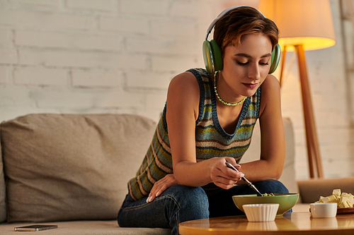 young woman in headphones eating vegetarian salad near tofu cheese while sitting on sofa in evening