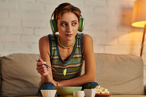 dreamy woman in headphones looking away while having evening vegetarian snack on couch at home