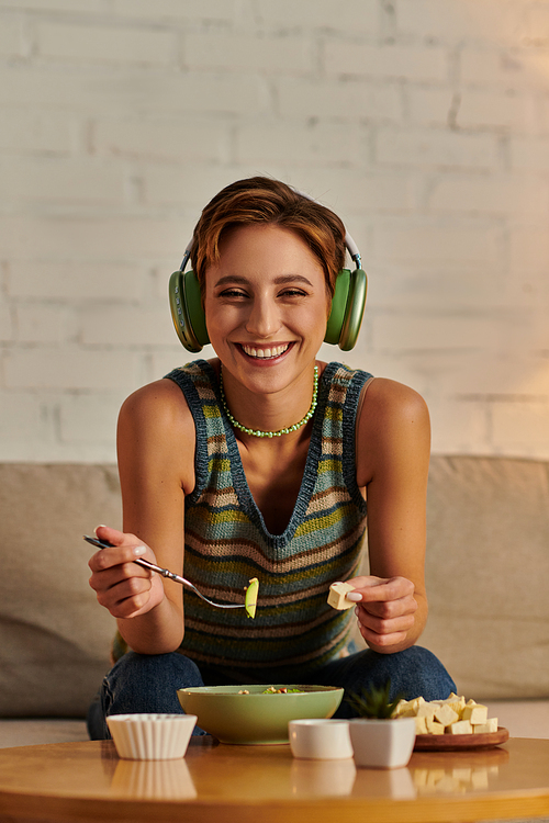 cheerful veggie woman in headphones looking at camera near plant-based salad and tofu cheese