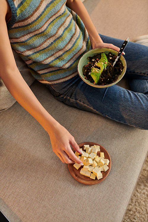 cropped view of woman with vegetarian salad sitting on couch near tofu cheese, evening snack