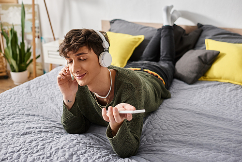 happy curly transgender person in wireless headphones holding smartphone in bedroom, listen music