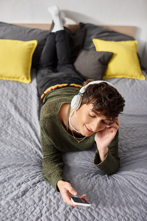 happy curly transgender person in wireless headphones holding smartphone in bedroom, listen music