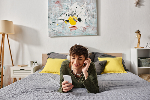 happy curly transgender person in wireless headphones holding smartphone in bedroom, listen music