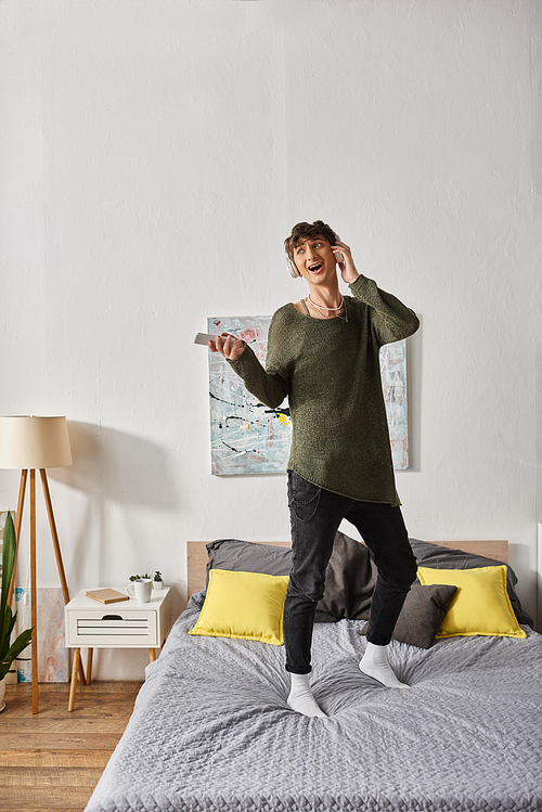 happy curly transgender person in wireless headphones holding smartphone and standing on bed