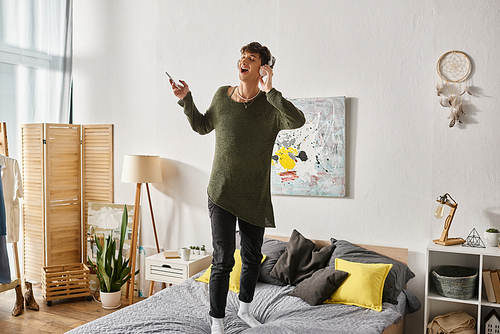 happy curly transgender person in wireless headphones holding smartphone and standing on bed