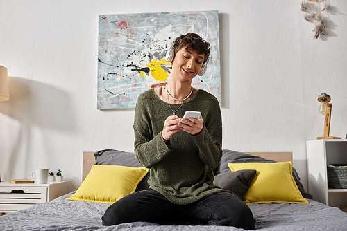 happy curly transgender person in wireless headphones holding smartphone and sitting on bed